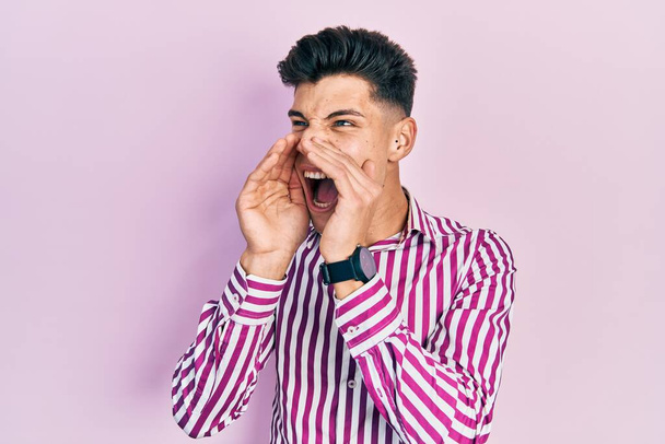jonge latino man dragen casual kleding schreeuwen boos hardop met handen over de mond  - Foto, afbeelding