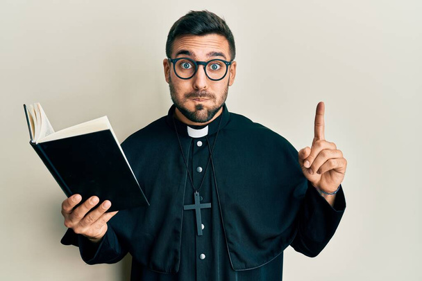 Молодий іспаномовний священик тримає Біблію пальцем, набиваючи щоки смішним обличчям. рот, надутий повітрям, ловить повітря
.  - Фото, зображення