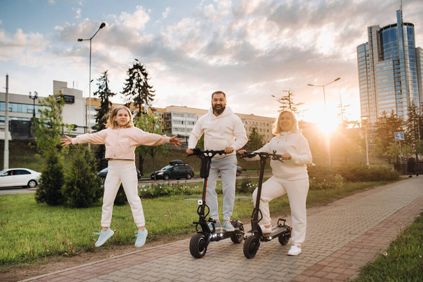 Une famille en vêtements blancs se tient dans la ville sur des scooters électriques au coucher du soleil. - Photo, image