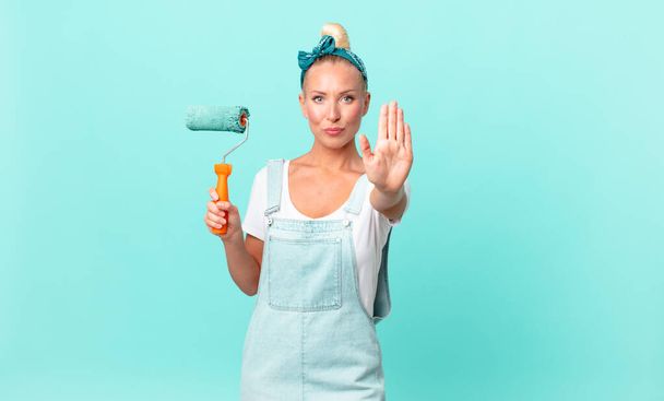 pretty blond woman looking serious showing open palm making stop gesture and painting a wall - Фото, изображение