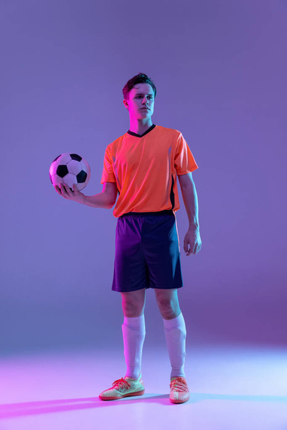 Full-length portrait of professional football player standing isolated on gradient blue pink background in neon light. Concept of sport, beauty. - Фото, изображение
