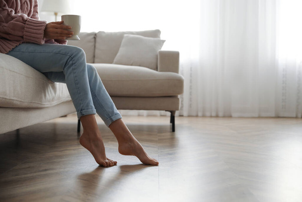Barefoot woman sitting on sofa in living room, closeup. Floor heating system - Fotografie, Obrázek