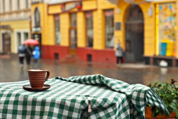 Coffee in a clay mug on a checkered tablecloth. Terrace of a street cafe on a background of blurred street. Sharpness on the mug. People with umbrellas on the background. - Foto, Bild