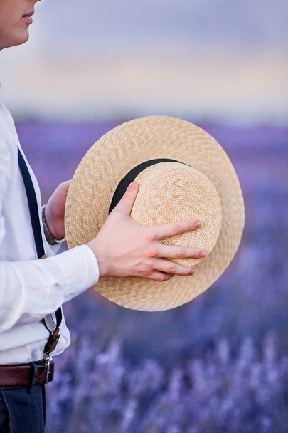 Young man, a groom in a white shirt, holds a straw hat in his hands close-up. Vertical photo while traveling in France - 写真・画像
