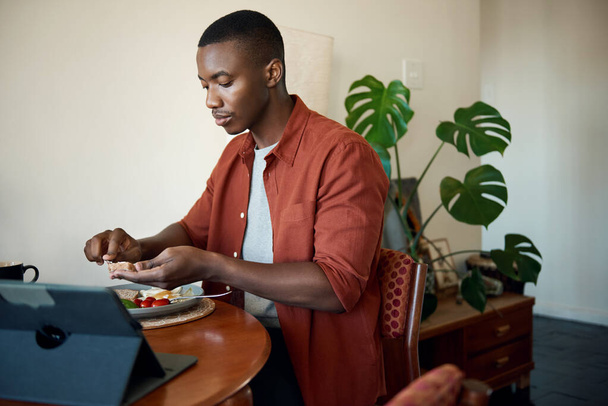 Young African man sitting at his dining room table enjoying a healthy breakfast and watching something on a tablet - 写真・画像