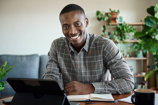Portrait of a smiling young African businessman working remotely from home at table in his living room using a digital tablet - Foto, Imagem