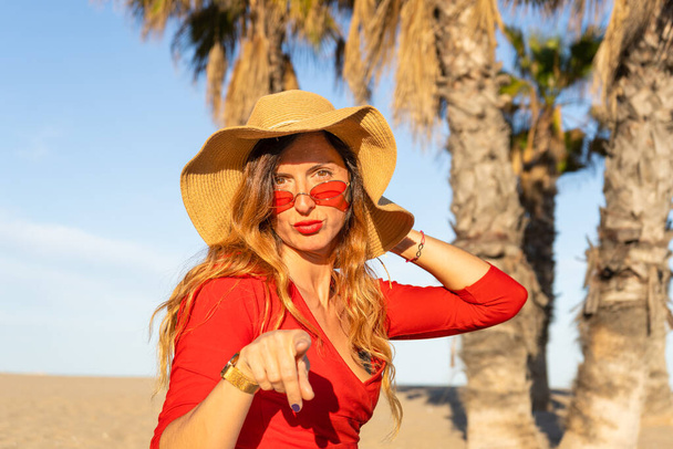 Donna che indica la fotocamera indossando cappello da sole, occhiali da sole rossi e camicia rossa su sfondo palmo della spiaggia. - Foto, immagini