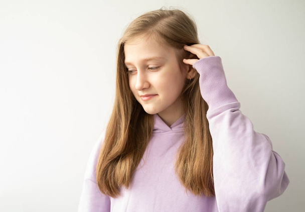beautiful teenage girl with long hair in purple huddyhoodie touching her hair standing against white wall - Photo, Image
