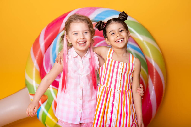 two happy little girls in colorful dress laughing hugging having fun on yellow background with lollipop - Photo, Image