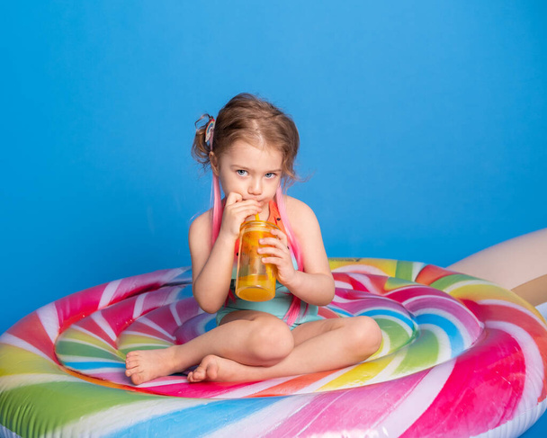 cute child girl in swimming suit drinking orange juice sitting on colorful inflatable mattress on blue background. - Photo, Image