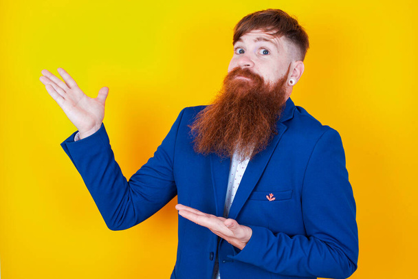 смущенный молодой бородатый мужчина в синей куртке позирует на желтом фоне - Фото, изображение