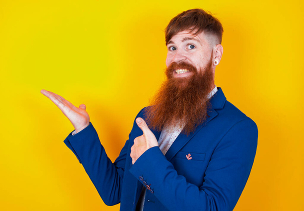молодой бородатый мужчина в синей куртке показывает большой палец вверх на желтом фоне - Фото, изображение