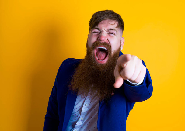 young bearded man wearing blue  jacket pointing to camera on a yellow background - Photo, Image