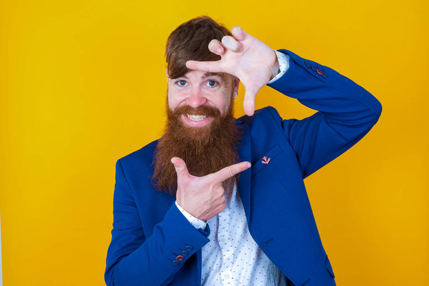 青いジャケットを着た若い髭の男が手で指のフレームを作ってる。創造性と写真の概念 - 写真・画像