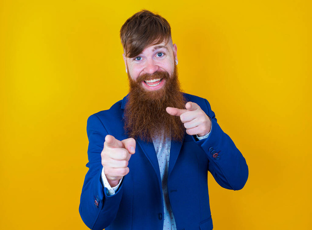 fiatal szakállas férfi kék kabátot visel, ami a kamera felé mutat sárga alapon - Fotó, kép