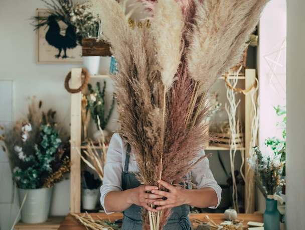 Nierozpoznawalna kobieta trzymająca Pampas w rękach w studio z suchymi kwiatami i roślinamiMłoda kobieta-kwiaciarnia stojąca z trawą pampas w rękach w kwiaciarni, koncepcja zrównoważonego stylu życia.  - Zdjęcie, obraz