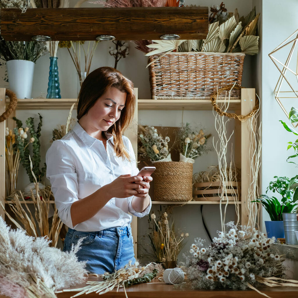 Cheerful Female Florist Using Mobile Phone in her Workshop Full With Different Dry Flowers and Herbs.Young talented smiling woman making everlasting dried plants and flowers arrangement in her flower shop and taking photo of them on her smartphone. - Photo, Image