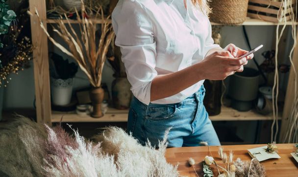 Unrecognizable Woman Entrepreneur Using Mobile Phone in her Workshop Full With Different Dry Flowers. Female florist making everlasting dried plants and flowers arrangement in her flower shop table, taking break to send text message on her smartphone - Foto, Bild