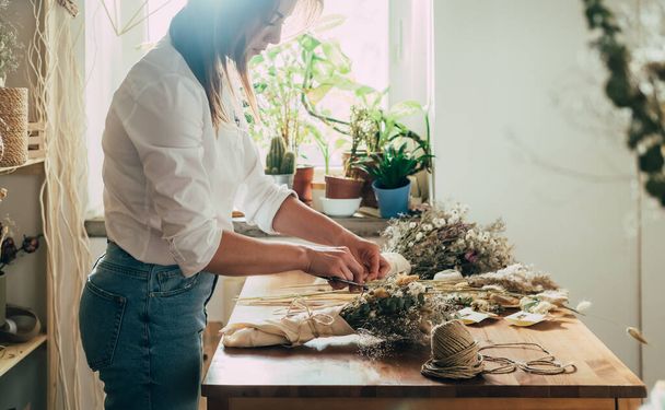 Beautiful Florist Arranging Bouquet of a Dry Flowers at her WorkshopSide view of young talented woman cutting with scissors dried plants and flowers arrangement in her flower shop, sustainable lifestyle concept.  - Foto, Imagen