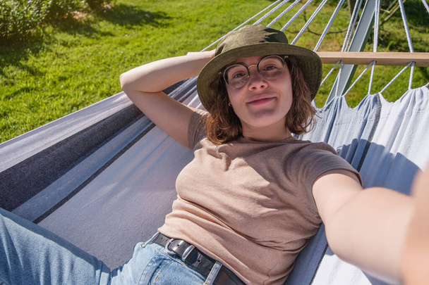 Autoportrait de jeune femme espagnole avec chapeau seau et lunettes couché dans hamac à l'extérieur. - Photo, image