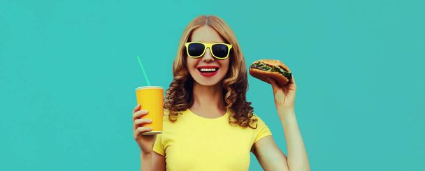Portrait of happy smiling young woman with fast food, burger and cup of juice wearing a yellow t-shirt, sunglasses on a blue background - Photo, image