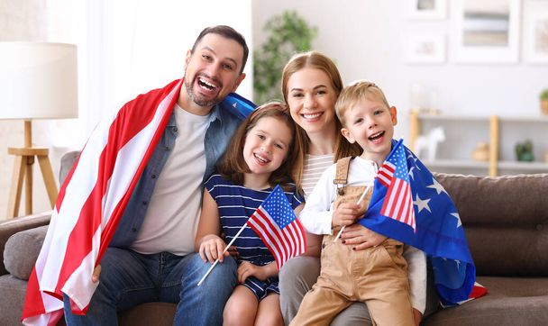 Nuoret onnelliset amerikkalaiset perheen vanhemmat ja kaksi pientä lasta istumassa sohvalla kotona Yhdysvaltojen lippujen kanssa ja hymyilemässä kameralle itsenäisyyspäivää vietettäessä. Isänmaallinen Yhdysvaltain loma käsite - Valokuva, kuva