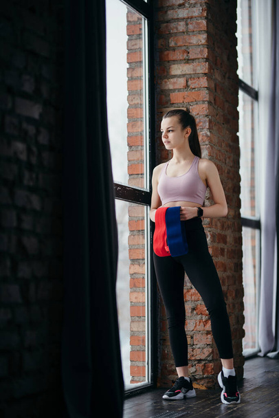 Beautiful young girl in a top and leggings holds elastic bands for workouts in her hands while standing in the gym - Zdjęcie, obraz