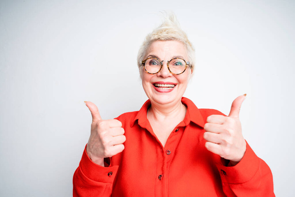 Happy elderly woman with glasses smiling and raising thumbs up while looking at the camera - Photo, Image