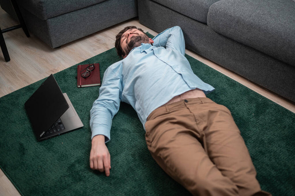 Jovem trabalhador de escritório bonito encontra-se no tapete de chão ao lado do sofá, ele está cansado e dormindo, vista superior - Foto, Imagem