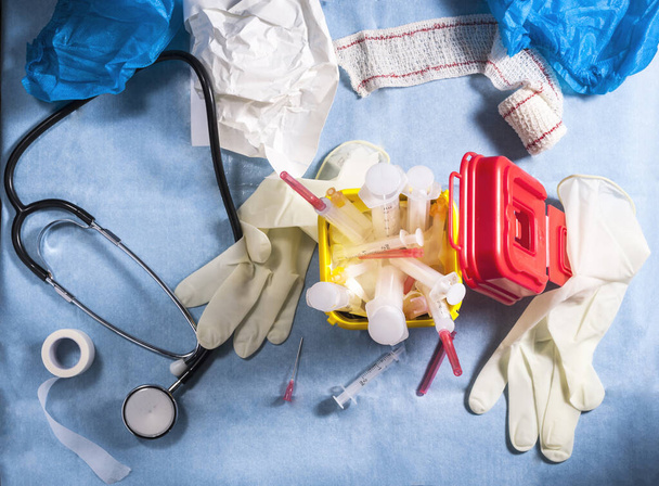 Aerial view of a sanitary still life in which you can see a yellow biological container full of syringes and needles and surrounded by bandages, a phonendoscope and surgical gloves - 写真・画像