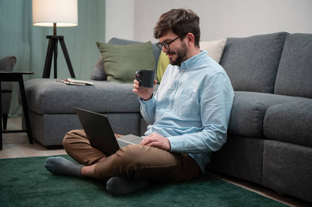 Durante o isolamento na quarentena do coronavírus, o cara se senta no chão, trabalha remotamente em um laptop e bebe de uma caneca de café. - Foto, Imagem