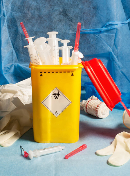 Still life of sanitary material.frontal view of a sanitary still life with a yellow biological container full of syringes and needles and around clinical material. behind a blue background of a surgical gown - Foto, imagen