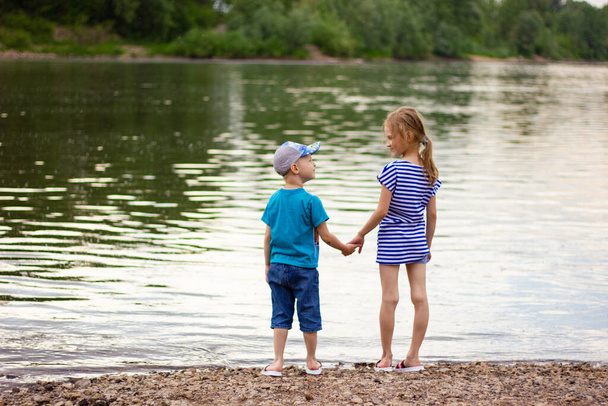 broer en zus houden elkaars hand vast en staan op de oever van de rivier op een zomeravond, kinderen vriendschap - Foto, afbeelding