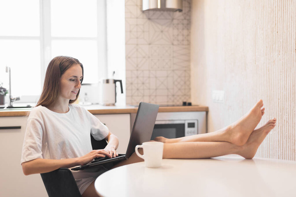 Jovem mulher trabalhando em laptop na cozinha. Freelancer feminino trabalhando online em casa. Foco seletivo - Foto, Imagem