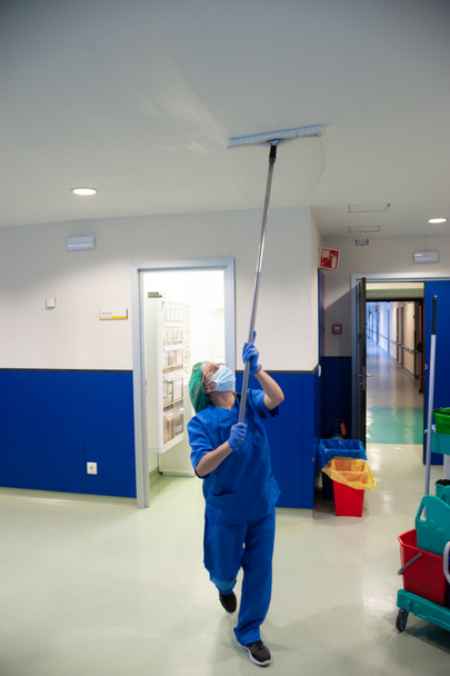 Mujer del servicio de limpieza de un hospital limpiando las ventanas de la sala de preoperatorio con un cepillo especial. Concepto sanitario - Foto, imagen