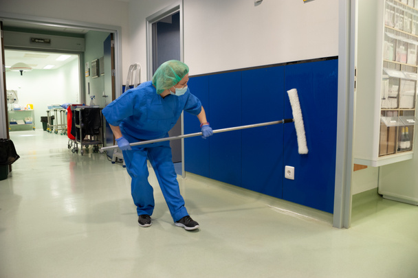 Mujer del servicio de limpieza de un hospital limpiando las ventanas de la sala de preoperatorio con un cepillo especial. Concepto sanitario - Foto, Imagen