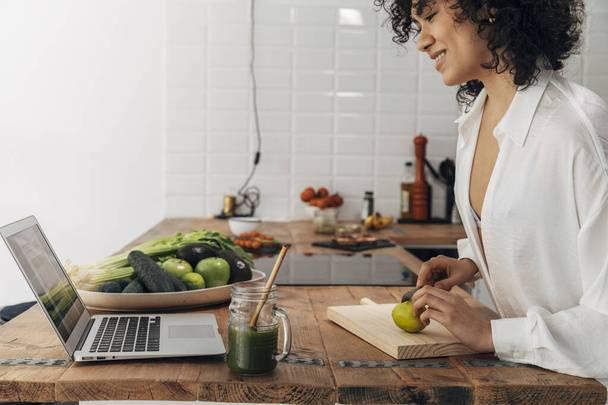 Smiling Young mixed race woman looking recipe laptop. Cutting lime with knife. Kitchen counter top. Green juice reusable straw. Technology concept. Health concept. Home concept. Copy space - Fotografie, Obrázek