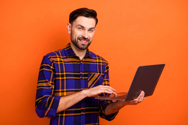Фото веселого молодого человека руки держать использовать нетбук зубастый улыбка смотреть камеры изолированы на оранжевый цвет фона - Фото, изображение