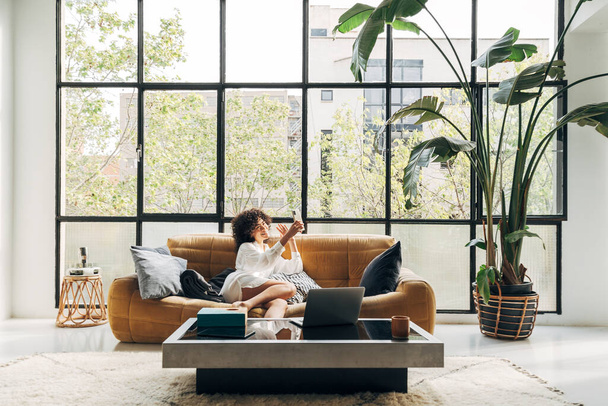Young African American woman sitting on a couch holding cellphone waving hand video calling distance friend. Home concept. Technology concept. Copy space - Foto, Bild