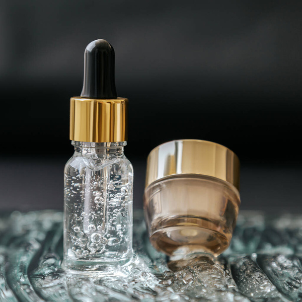 Skincare cosmetic Serum bottle with fluid collagen hyaluronic acid, moisturizer cream jar on water gel bubbles texture surface. Luxury gold water balance cosmetic on black background. Square Close up - Fotografie, Obrázek