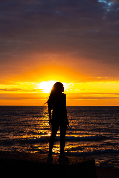 A beautiful African American woman sunrise silhouette on Lake Michigan as she looks out across the water with the orange and yellow clouds reflecting on the water below. - Foto, imagen