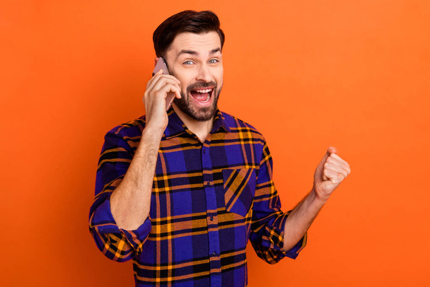 Photo of astonished man speak phone open mouth fist up celebrate wear plaid isolated on orange color background - Foto, Imagem