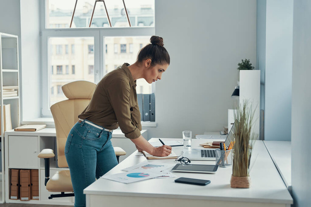 Beautiful young woman in smart casual wear writing something down while standing in the office - Foto, imagen