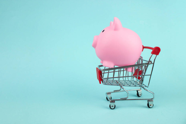 Pink piggy bank in mini shopping cart on blue background. Saving money for sales of the year, black friday concept - Photo, Image