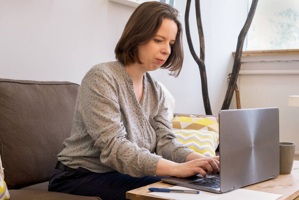 Young brunette woman with shoulder length hair concentrated working on laptop. Woman is sitting at home on the couch in front of a table with a laptop with a cup of coffee. Remote work from home - Fotoğraf, Görsel