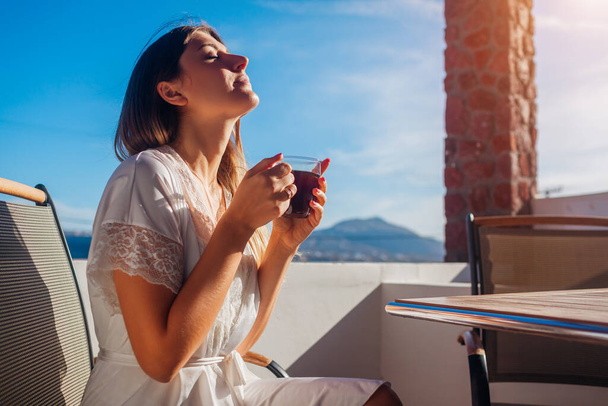 Γυναίκα χαλαρώνει στη βεράντα του ξενοδοχείου πίνοντας πρωινό καφέ απολαμβάνοντας το ορεινό τοπίο της Σαντορίνης στο Ακρωτήρι. Καλοκαιρινές διακοπές - Φωτογραφία, εικόνα