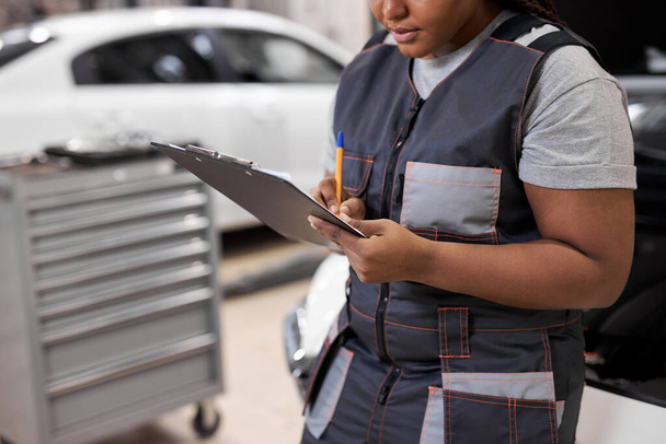 black female Auto Mechanic Making Car Checkup, in Uniform Writing and holding Clipboard - Photo, image