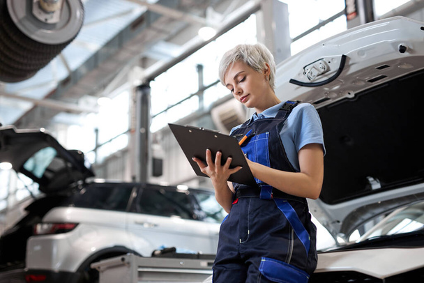 Side view on female Auto mechanic checking car engine, writing checklist on clipboard - Zdjęcie, obraz