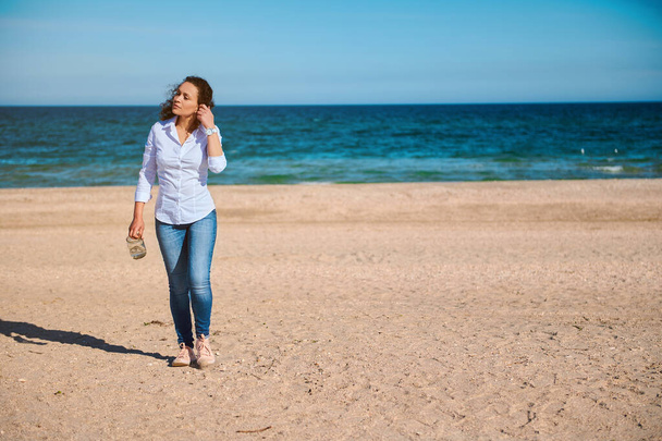 jong gemengd ras vrouw met krullend haar lopen op het strand op een zonnige dag - Foto, afbeelding