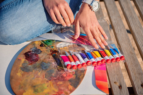 Close-up of female artist hands on a painting palette and tubes with paints on a wooden surface - 写真・画像